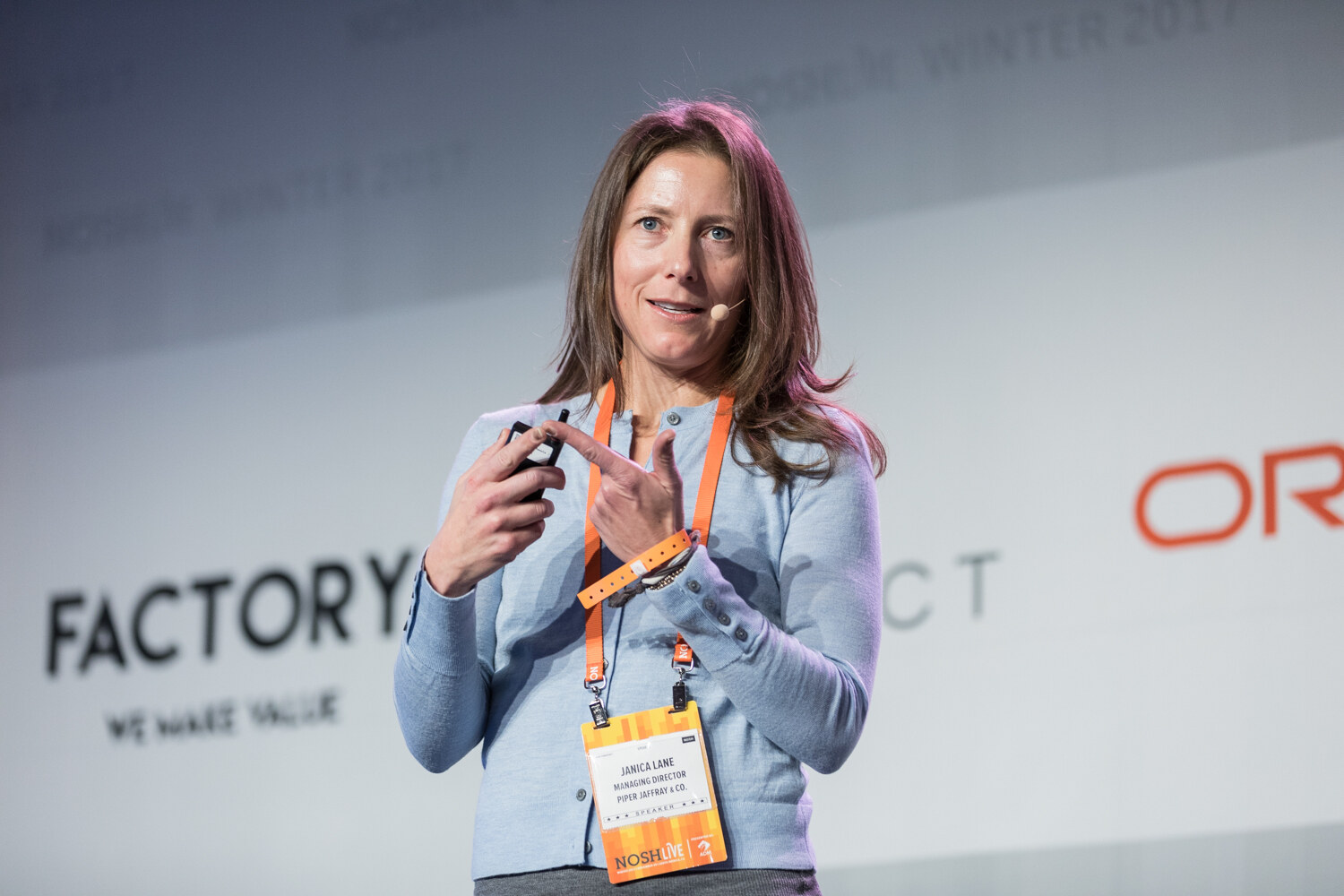 A woman with long brown hair and a headset microphone points with one hand while holding a slide clicker, in front of a Factory banner.