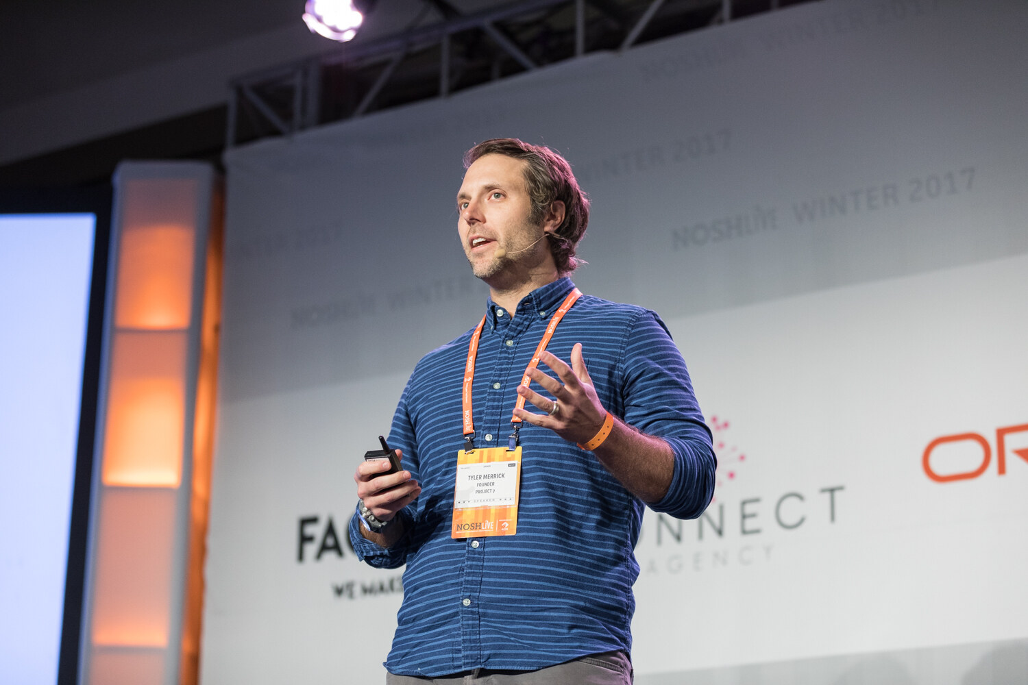 A man in a blue striped shirt with an orange lanyard and a slide clicker speaks on stage in front of Factory and Konnect banners.