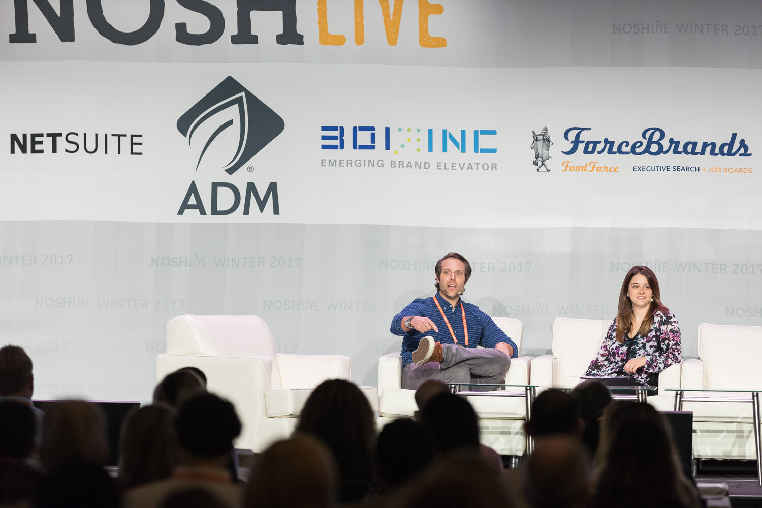 A man in a blue striped shirt gestures while seated next to a woman in a floral top on a stage beneath a NOSH Live sponsor banner, with audience heads in the foreground.