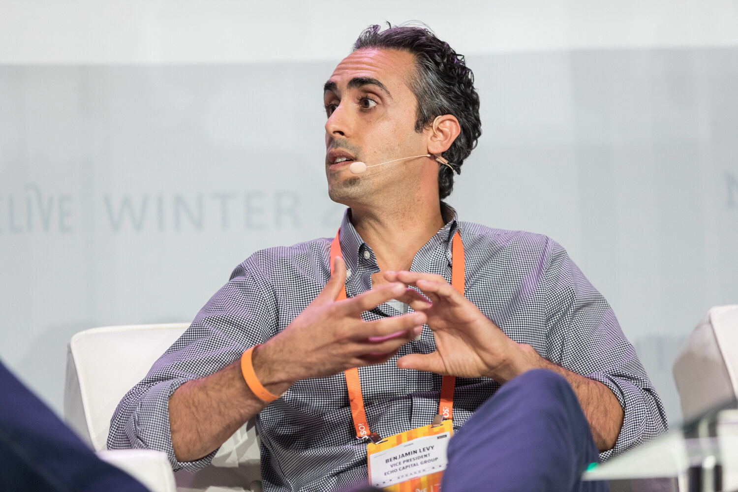 A dark-haired man in a gray checked shirt gestures with both hands while seated on stage, wearing an orange lanyard.