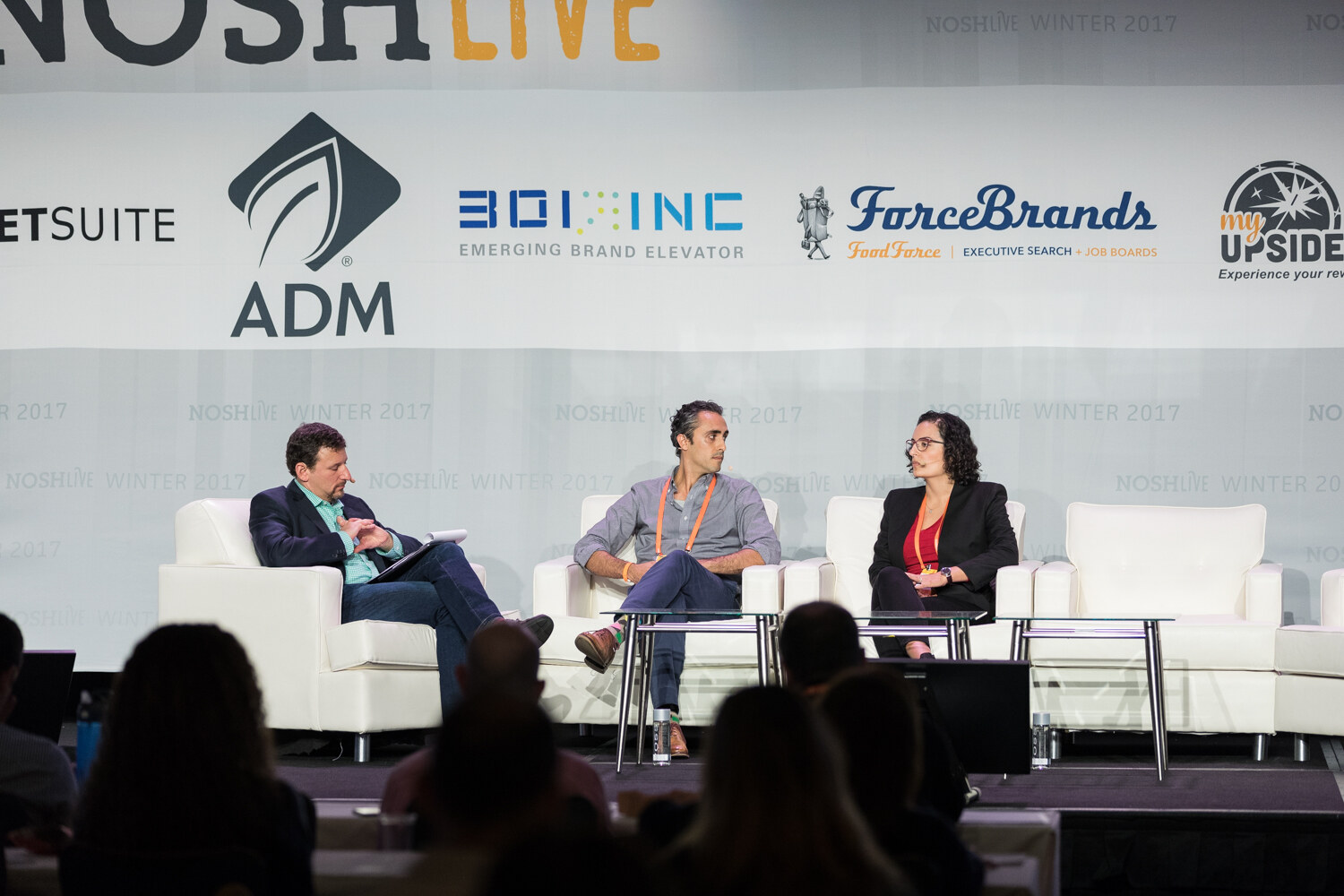 Three people sit in white armchairs on stage beneath a NOSH Live backdrop with ADM, 301 Inc, ForceBrands and My Upside logos, with audience heads silhouetted in front.
