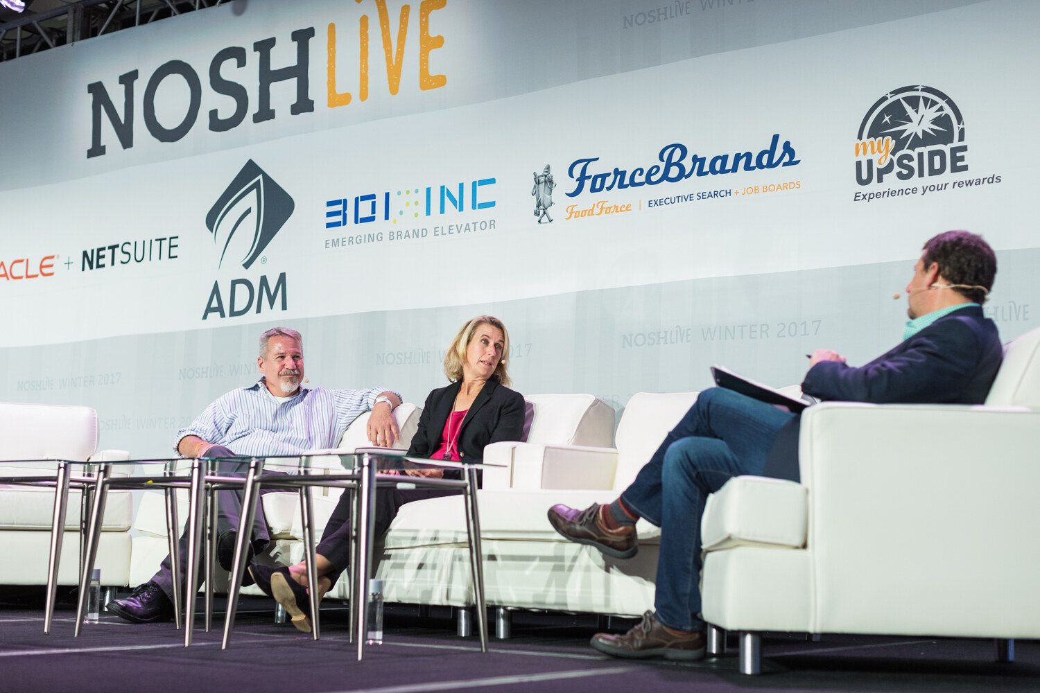 Two panelists and a moderator in white armchairs on a low stage under a NOSH Live sponsor banner.