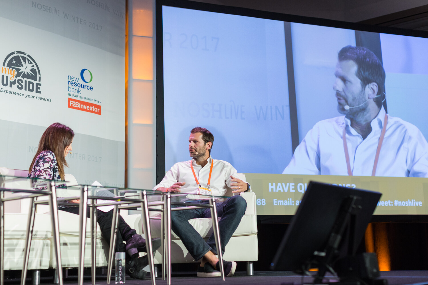 A woman seated at left faces a man in a white shirt on stage, with a large screen at right showing him in close-up and a Have Questions prompt below.