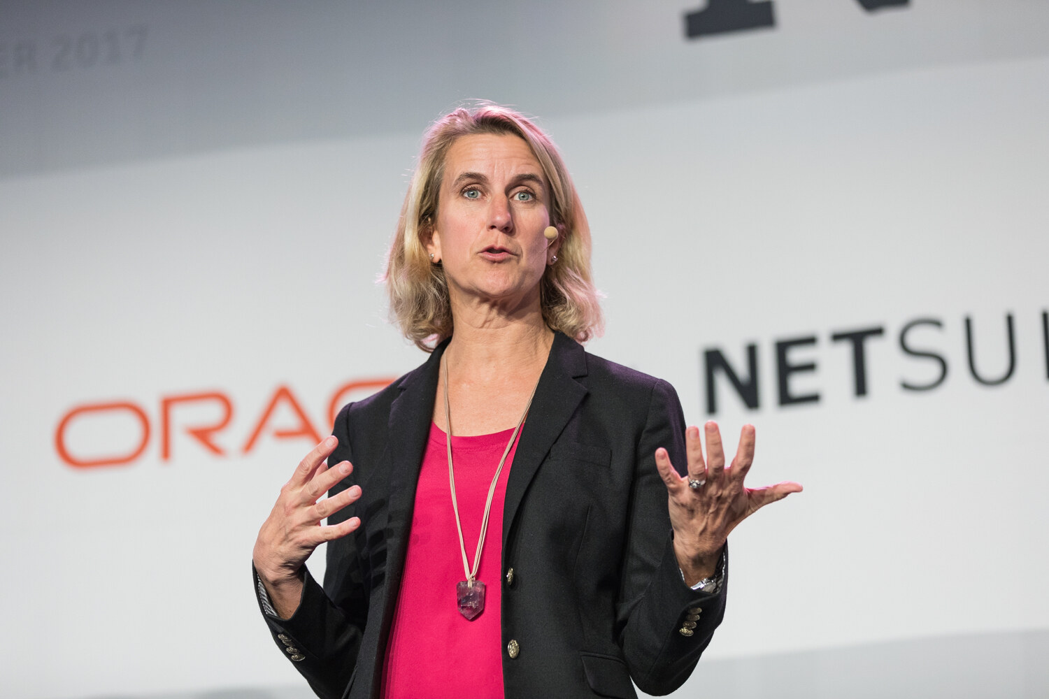 A blonde woman in a black blazer and pink top speaks with both hands raised in front of an Oracle NetSuite banner.