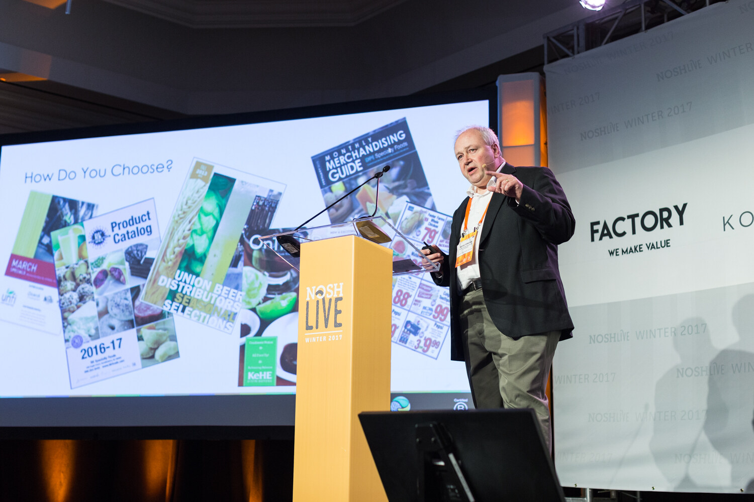 A man in a dark blazer at a yellow NOSH Live podium points at a projected slide titled How Do You Choose showing product catalogs and a merchandising guide.