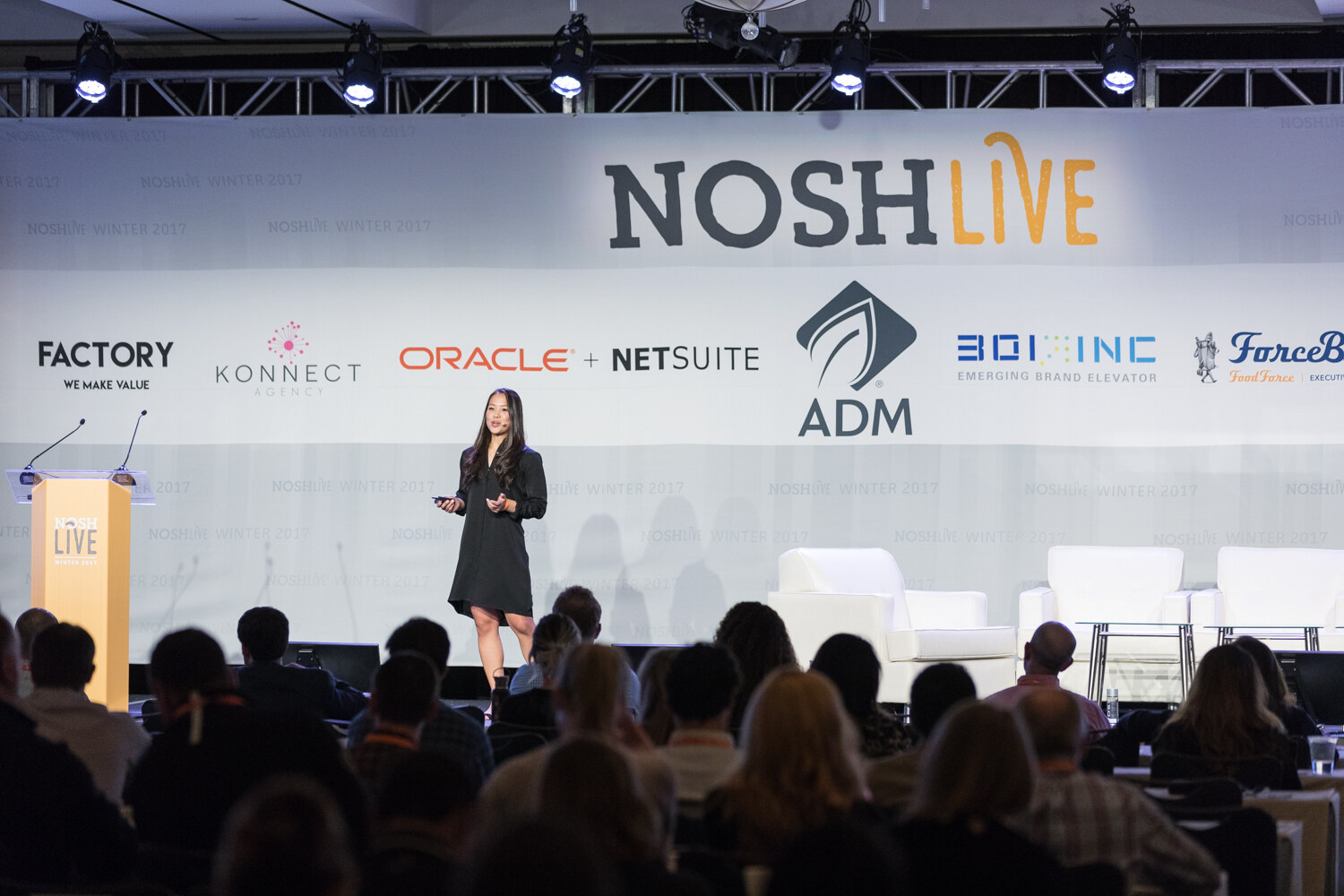 A woman in a black dress holds a clicker on a stage under a NOSH Live banner with Factory, Konnect, Oracle NetSuite, ADM and 301 Inc logos, with audience heads in the foreground.