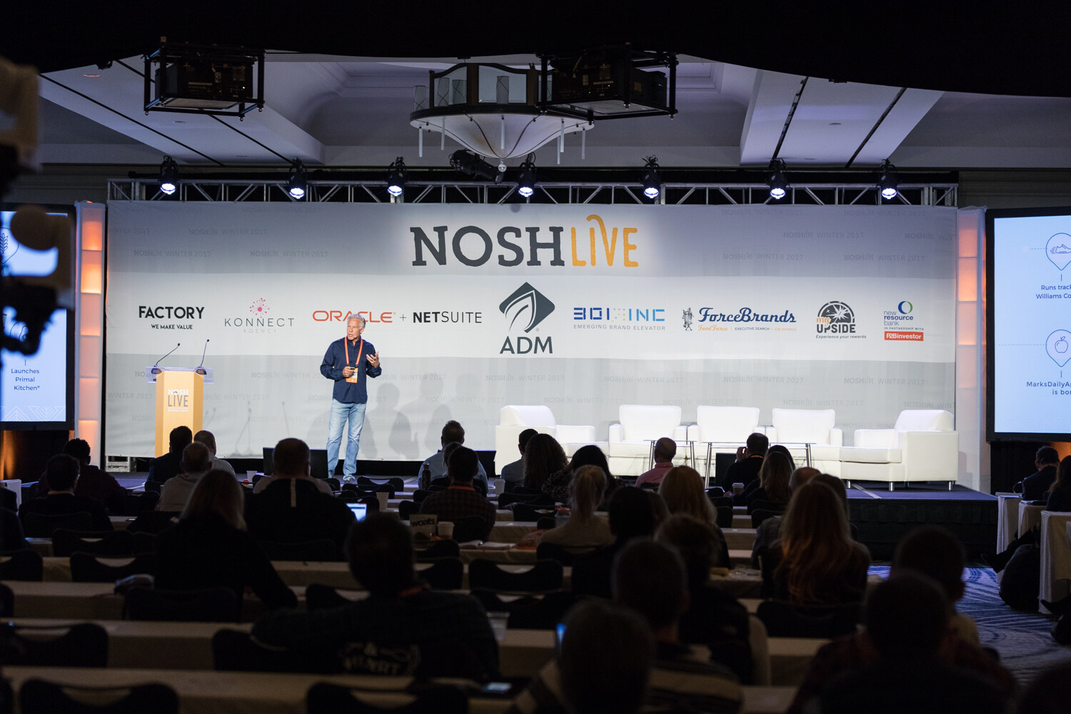 A wide view of a ballroom with a speaker on a stage under a NOSH Live sponsor backdrop, rows of seated attendees in the foreground and a camera at left.