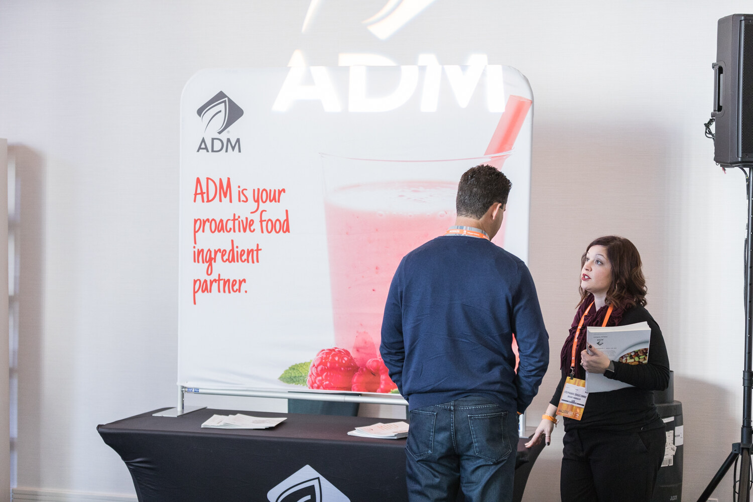 A man in a navy sweater stands facing a woman in black holding a booklet, beside an ADM banner reading ADM is your proactive food ingredient partner.
