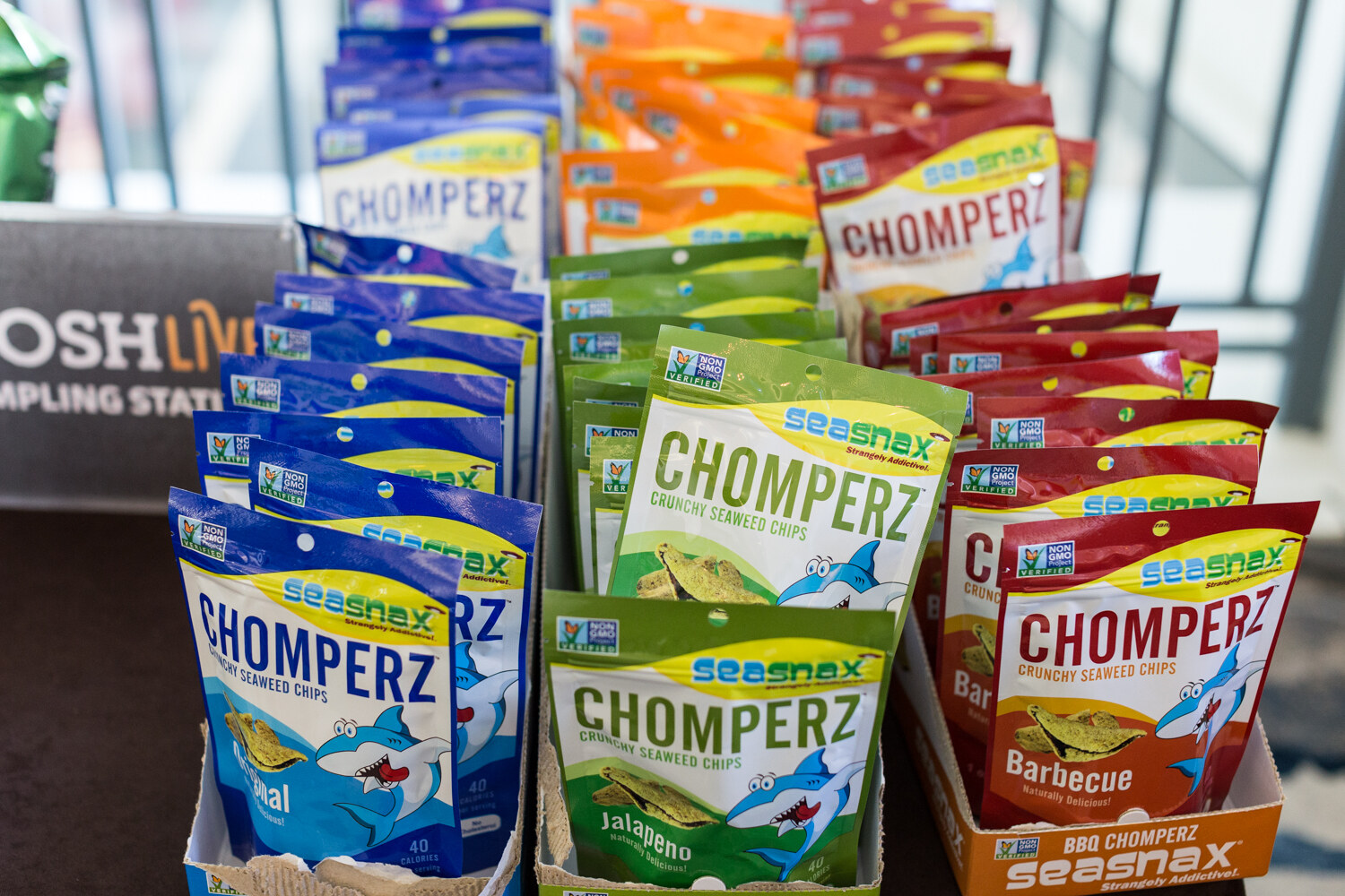 Rows of Chomperz crunchy seaweed chips bags in blue, green and red packaging, with a NOSH Live Sampling Station sign partly visible at left.
