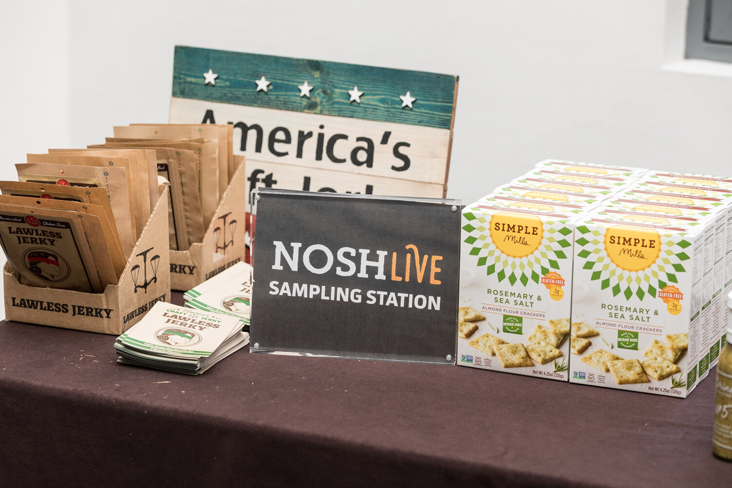 A table with a sign reading NOSH Live Sampling Station, beside Lawless Jerky packs in cardboard holders and boxes of Simple Mills almond flour crackers.