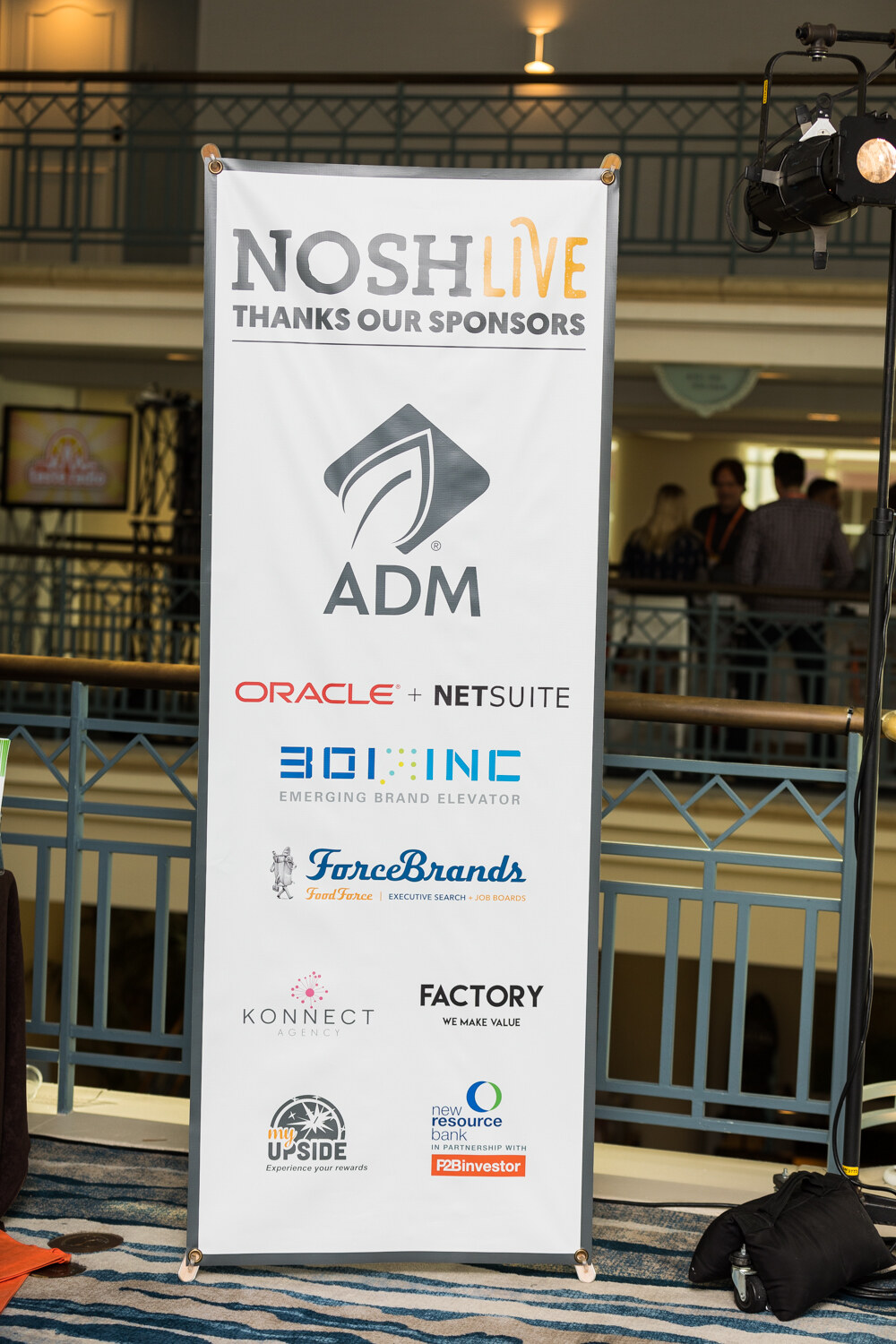 A tall banner reading NOSH Live Thanks Our Sponsors, listing sponsor logos, stands on a hotel balcony.