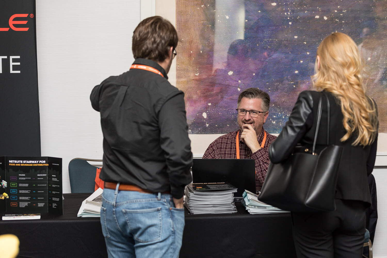 A seated man in a plaid shirt speaks with two attendees standing at a black-draped table with stacks of brochures and a banner showing part of the Oracle logo.