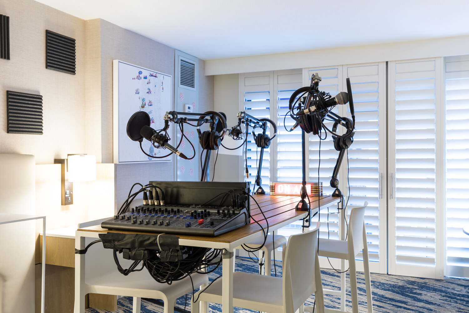 A hotel room with a wooden table holding an audio mixer, four boom microphones with headphones and an illuminated On Air sign, with white shutters behind.