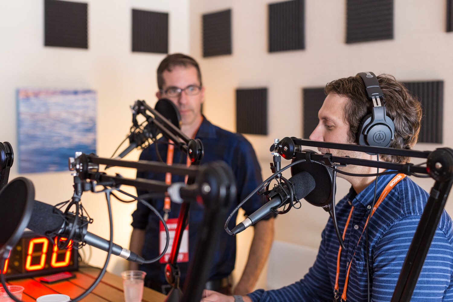 A man in a blue striped shirt and headphones sits at a boom microphone while a man with glasses stands behind a second microphone, beside a red digital timer reading 00.