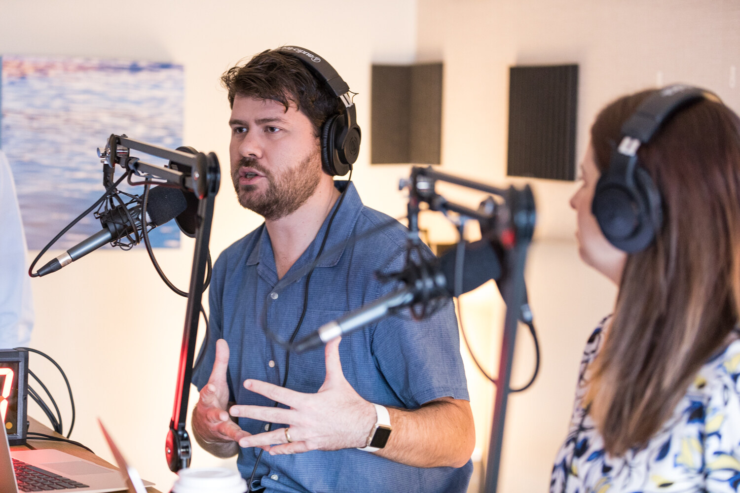 A bearded man in a blue shirt and headphones gestures while speaking at a boom microphone, with a woman in headphones beside him and a studio timer at left.