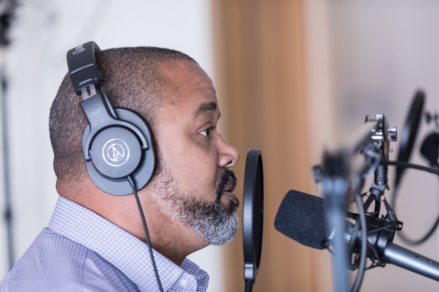 A bearded man in a lavender checked shirt and over-ear headphones speaks in profile into a condenser microphone with a pop filter.