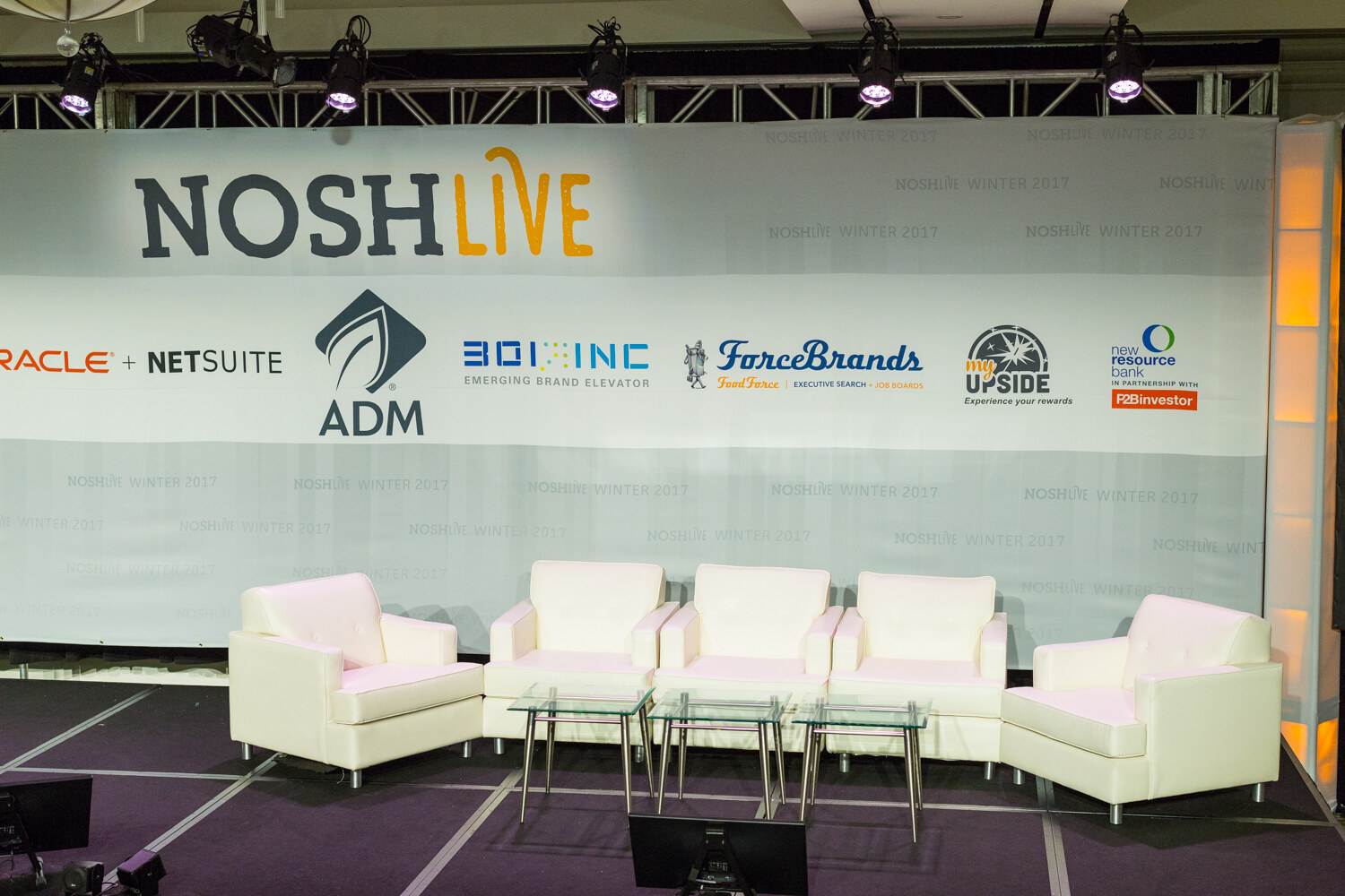 Five white armchairs and glass coffee tables on a purple-carpeted stage, below a NOSH Live banner with Oracle NetSuite, ADM, 301 Inc, ForceBrands, My Upside and New Resource Bank logos.