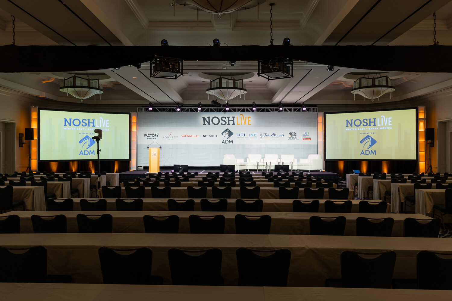A wide view of an empty ballroom with rows of tables and chairs facing a stage with a NOSH Live sponsor backdrop and a lectern, flanked by two screens showing the NOSH Live logo and ADM.
