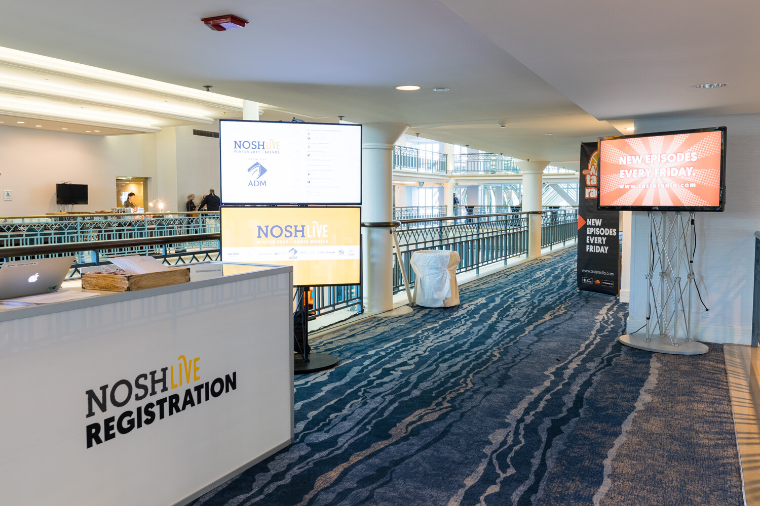 A white desk reading NOSH Live Registration beside two screens showing the NOSH Live agenda, a Taste Radio banner and a monitor reading New Episodes Every Friday, along a blue wave-patterned carpet.