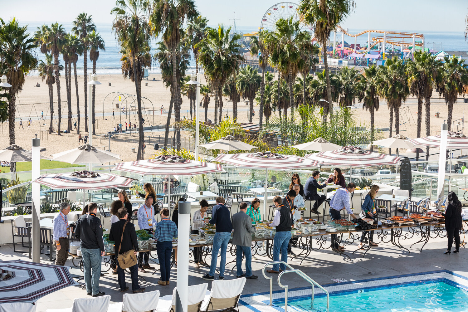 An elevated view of a hotel pool terrace with striped umbrellas and a lunch buffet, with a row of palm trees, a sandy beach and the Santa Monica Pier Ferris wheel behind.