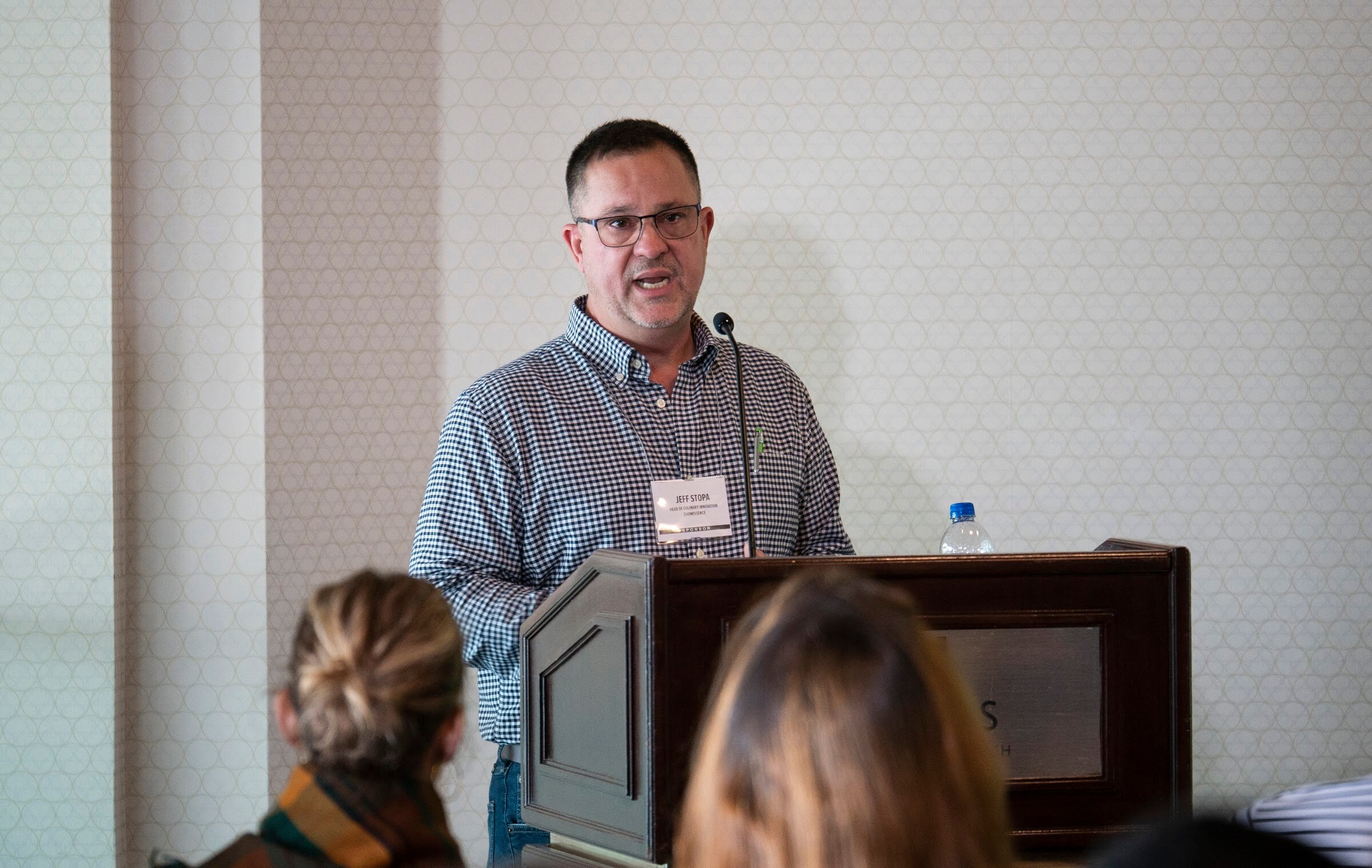 A man in a checked shirt speaks at a wooden lectern with a microphone.