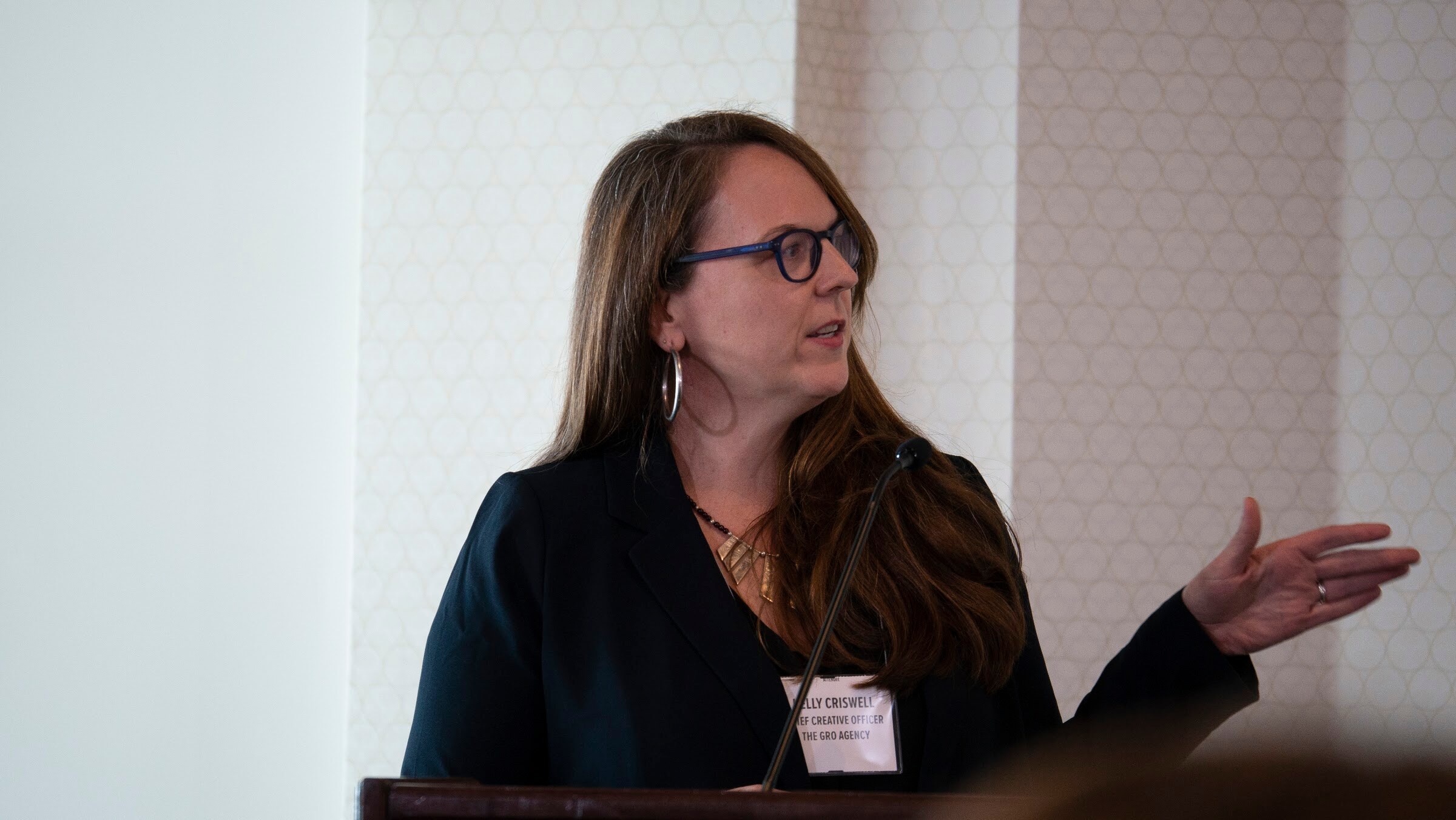A woman in glasses and a dark blazer gestures while speaking at the lectern.