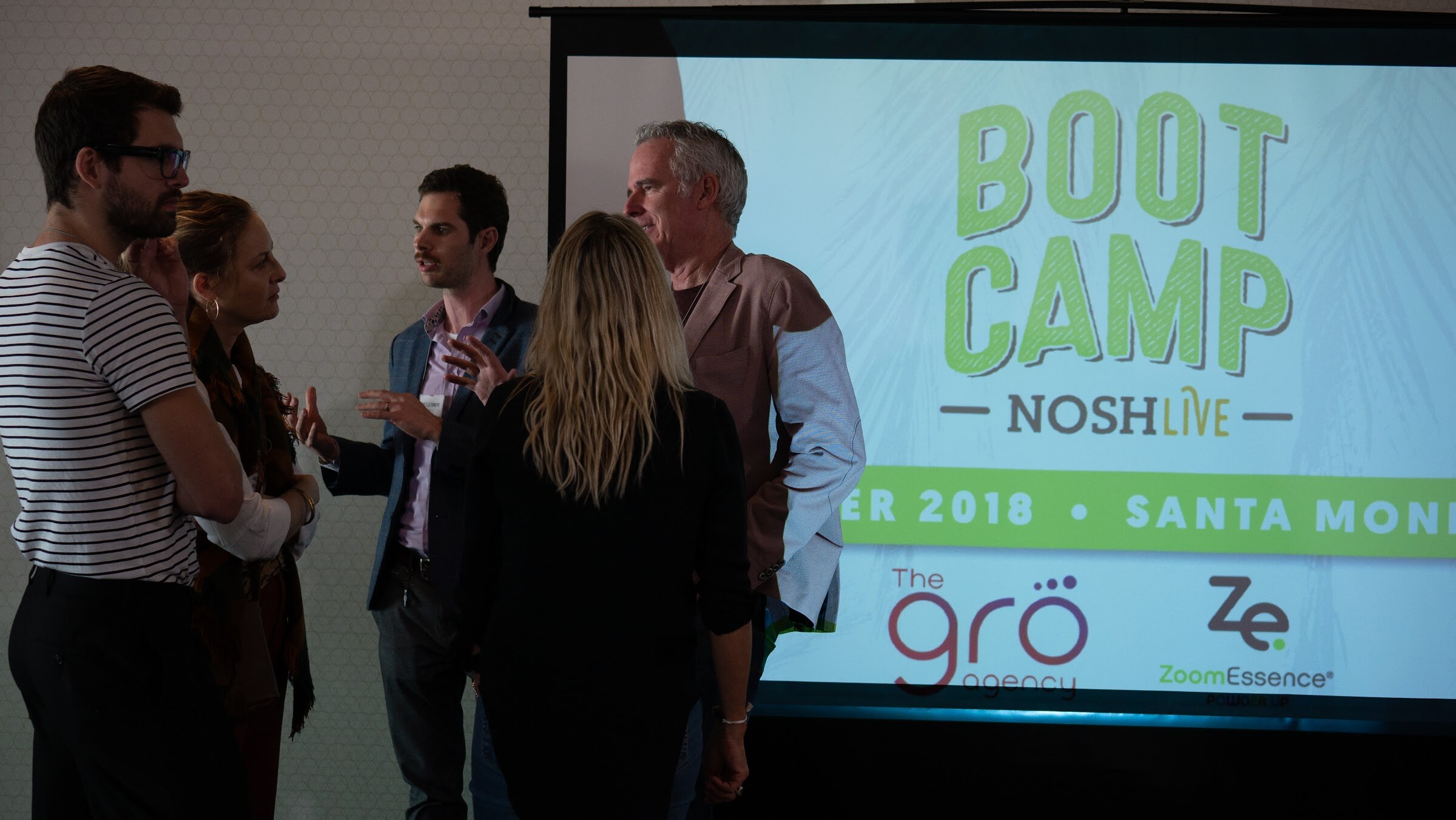 Attendees chat in front of a projected Boot Camp Nosh Live Winter 2018 Santa Monica slide.
