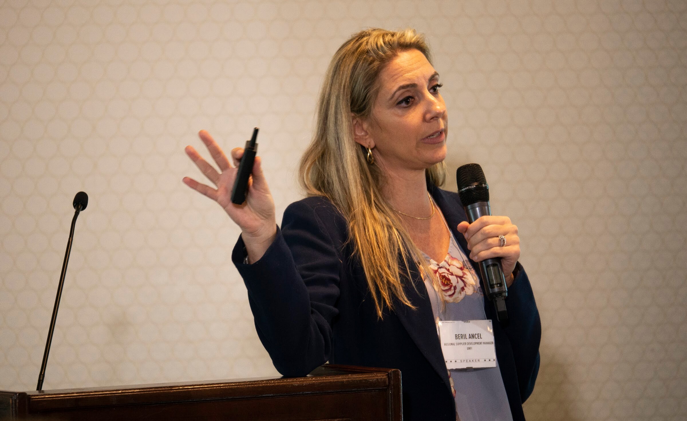A woman in a navy blazer holds a microphone and a clicker while presenting.