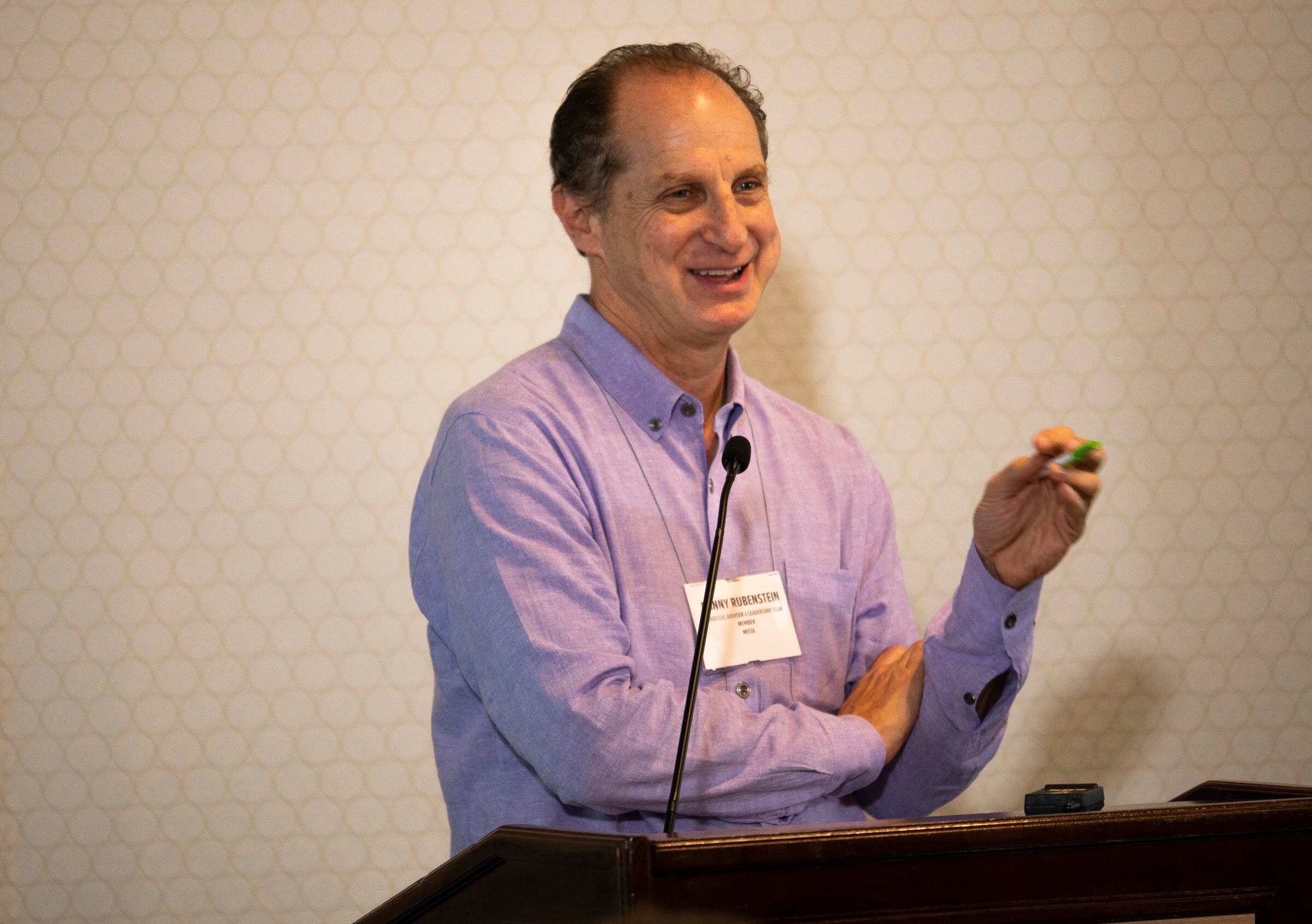 A man in a lavender shirt smiles and holds up a snack while presenting at the lectern.