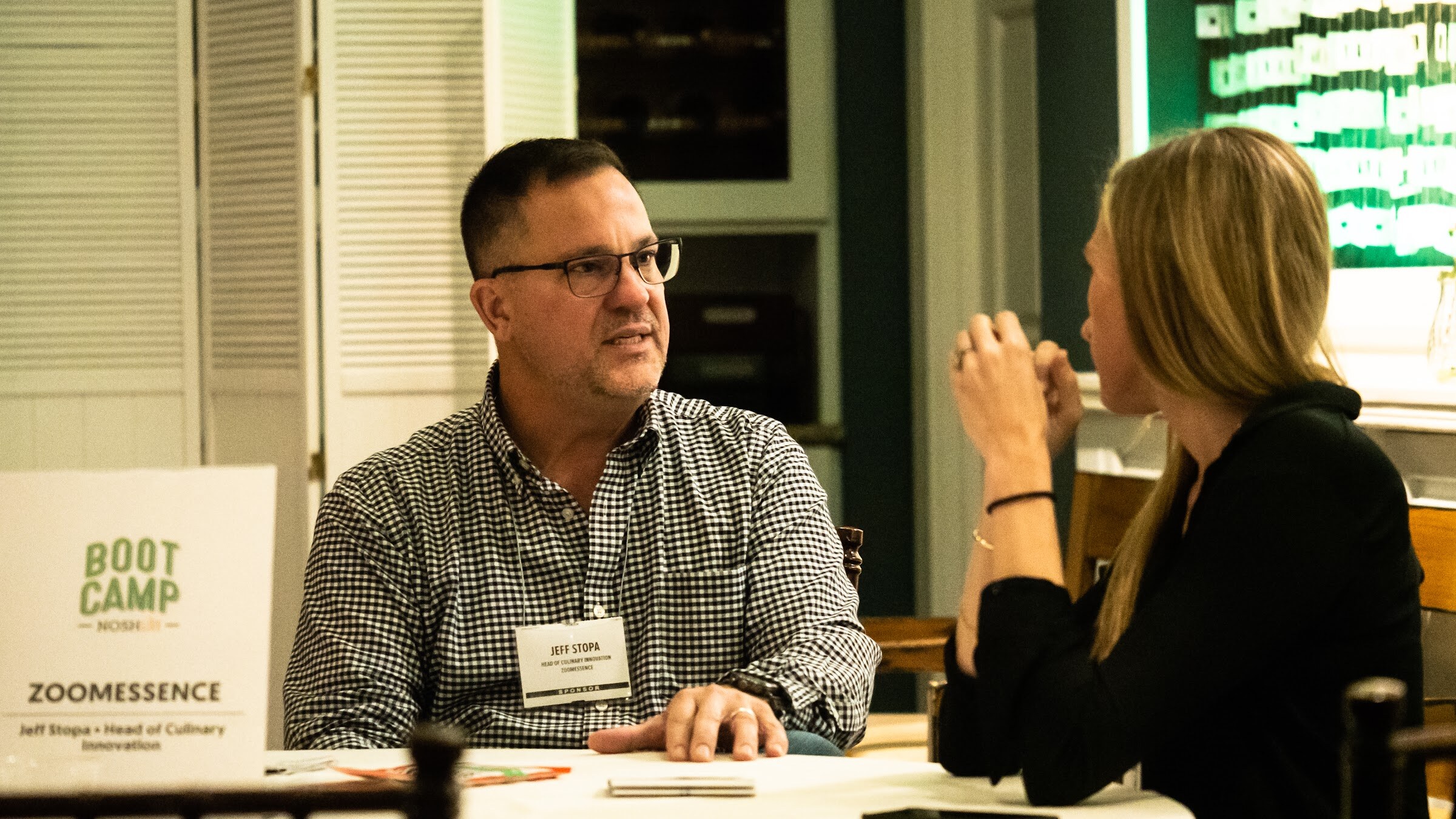 A man in a gingham shirt talks with a woman at a table with a Zoomessence card.
