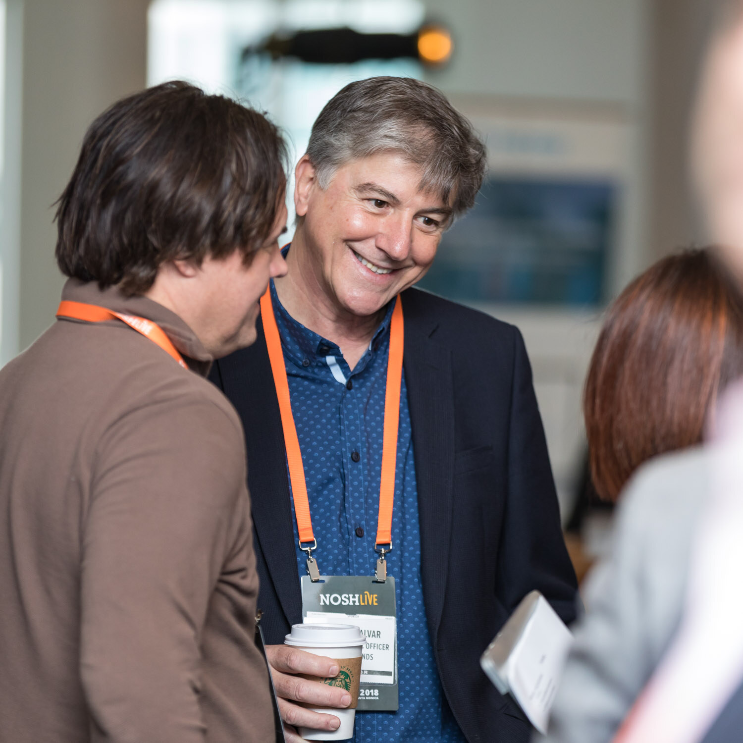 A smiling man wearing a Nosh Live badge talks with other attendees.