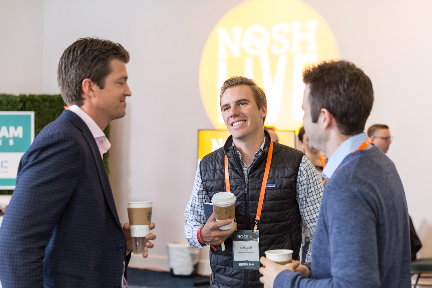 Two men talk and smile with coffee in front of a Nosh Live sign.