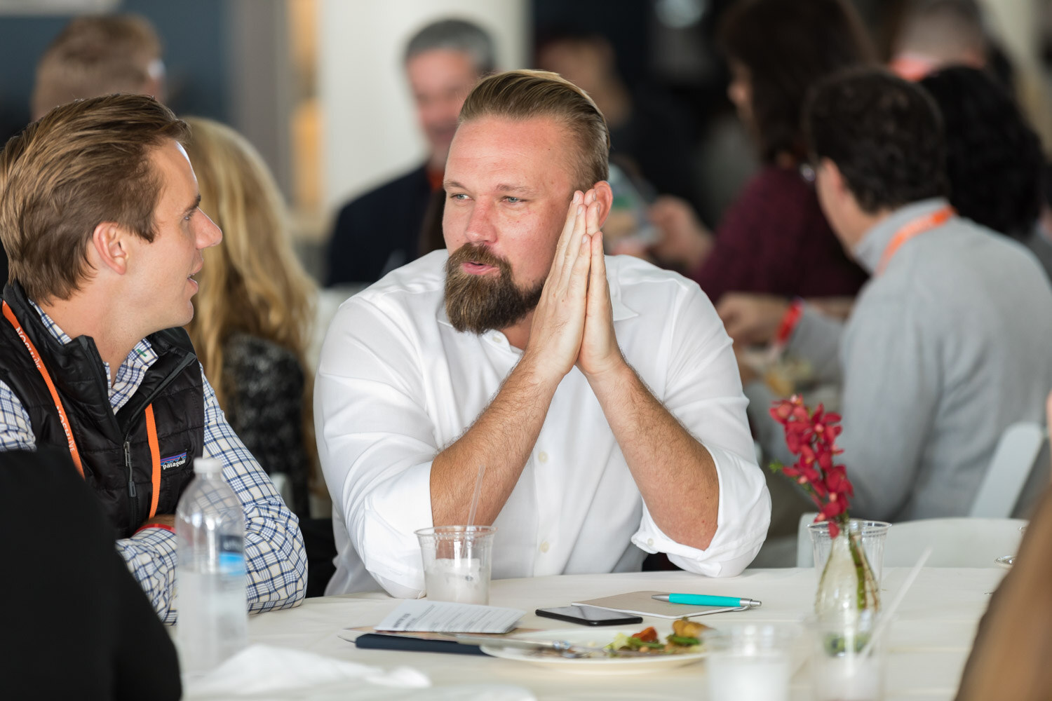A bearded man in a white shirt presses his hands together while listening at a lunch table.