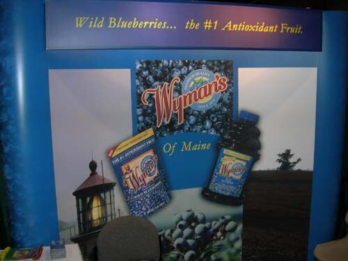 Blue Wyman's booth with a banner reading Wild Blueberries, the #1 Antioxidant Fruit, images of a lighthouse, a frozen blueberry bag and a juice bottle, and a chair in front.