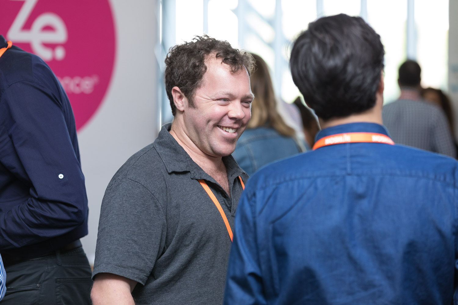 A bearded man laughs while talking with other attendees near a Zoomessence banner.