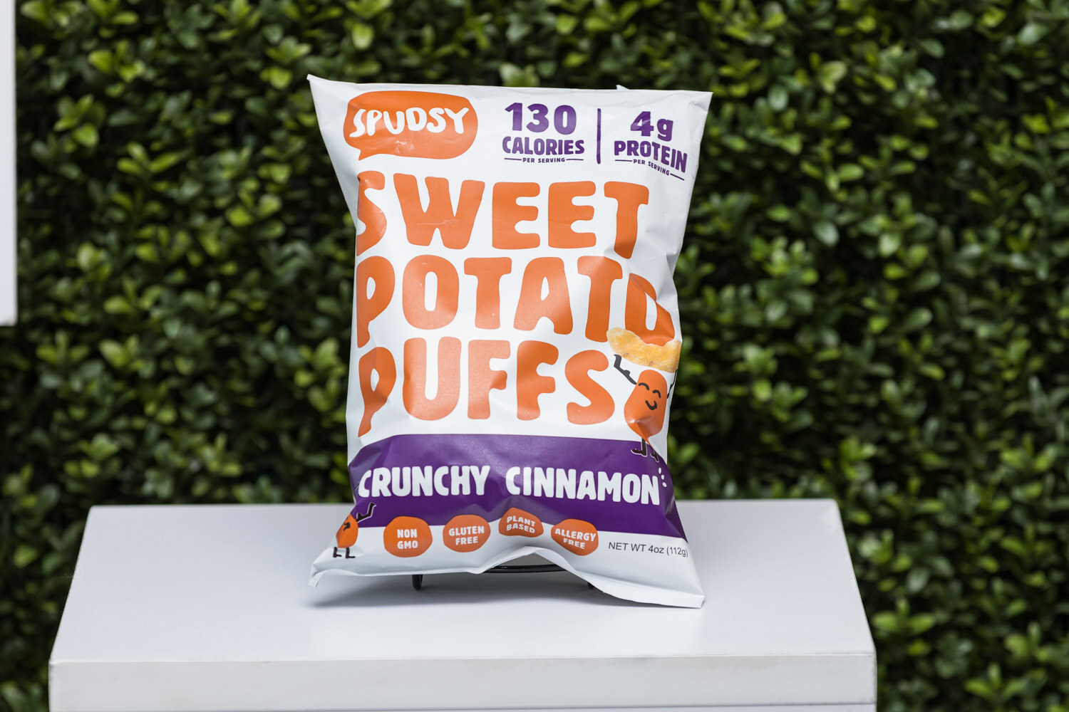 A bag of Spudsy Sweet Potato Puffs Crunchy Cinnamon on a pedestal in front of a hedge wall.