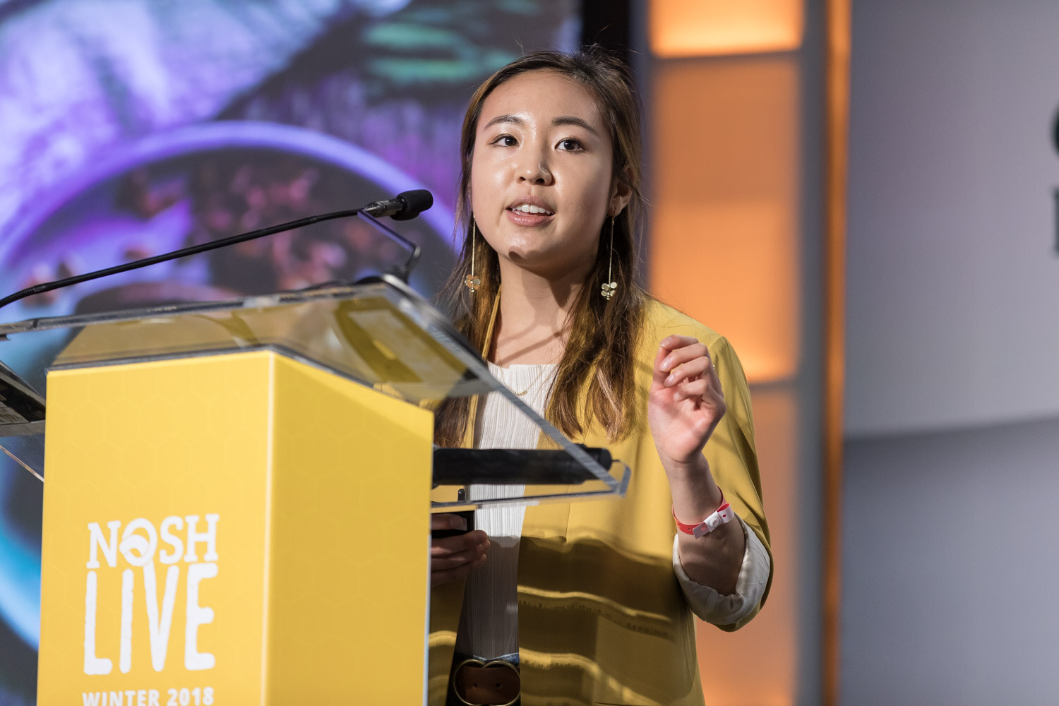 A woman in a yellow blazer pitches at the Nosh Live lectern with a colorful food photo on the screen.