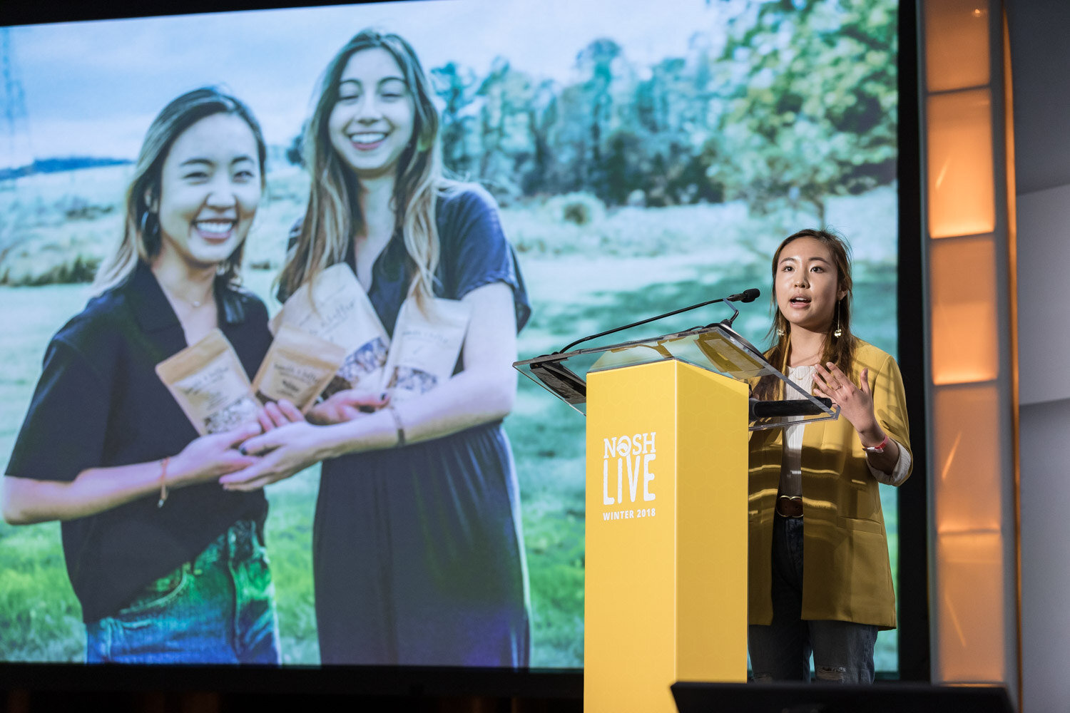A pitch slide shows two women holding packaged products while the presenter stands at the lectern.