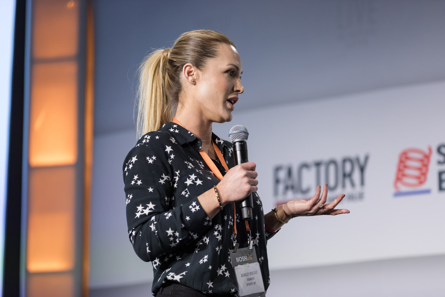 A woman in a star-print blouse holds a microphone and gestures while pitching.