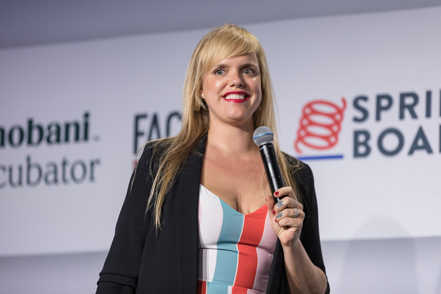 A woman in a black blazer and striped jumpsuit smiles while holding a microphone on stage.