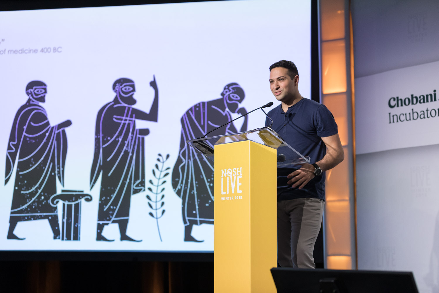 A presenter in a navy shirt stands at the lectern beside a slide of ancient Greek figures.