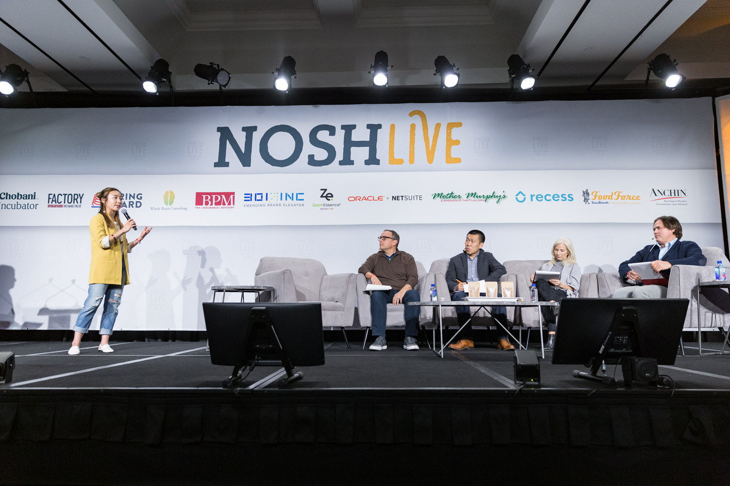 A pitch presenter in a yellow blazer holds a microphone on the main stage while four judges sit on couches behind her.