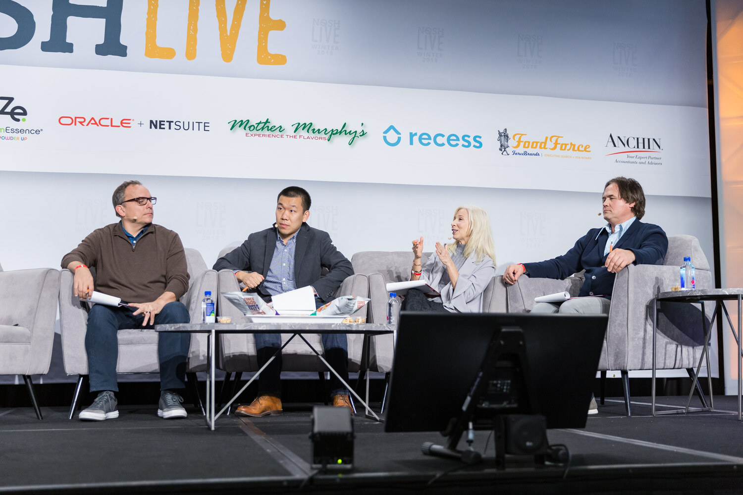 Four Pitch Slam judges sit on gray couches on the main stage with notepads and product samples.