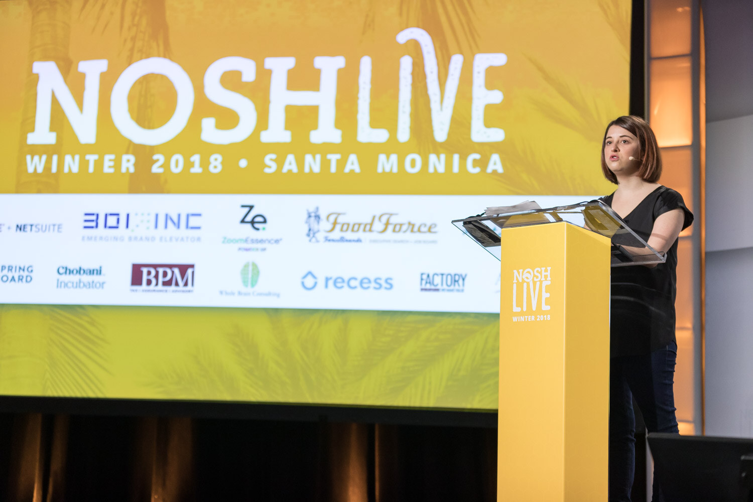 A woman in black speaks at a yellow Nosh Live lectern in front of the Nosh Live Winter 2018 Santa Monica screen with sponsor logos.