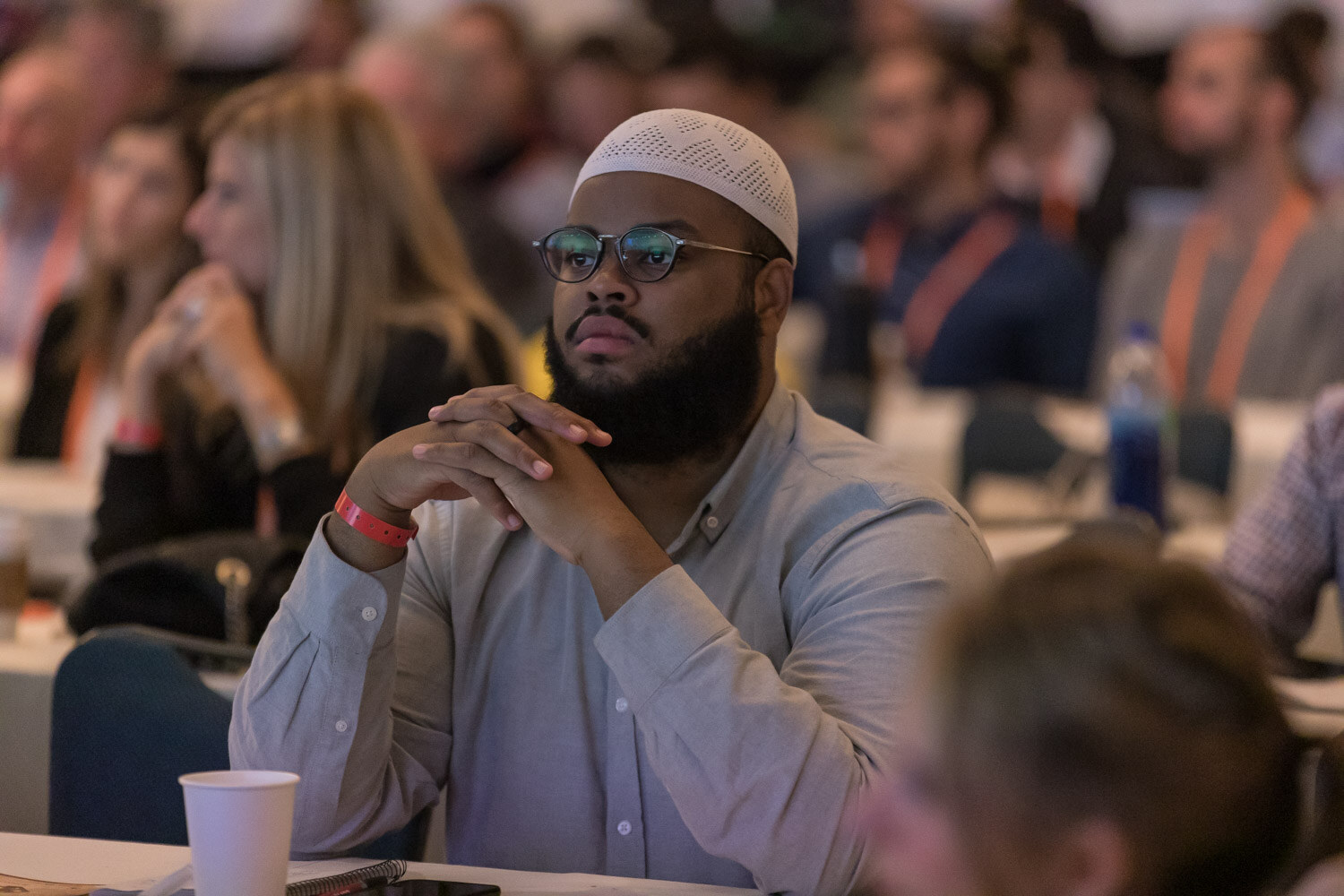 A bearded man in glasses and a white kufi listens with his hands steepled in the audience.