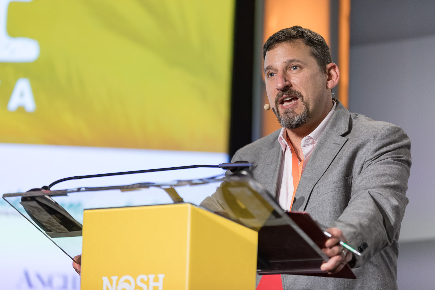 A man in a gray blazer with a headset microphone speaks at the Nosh Live lectern.