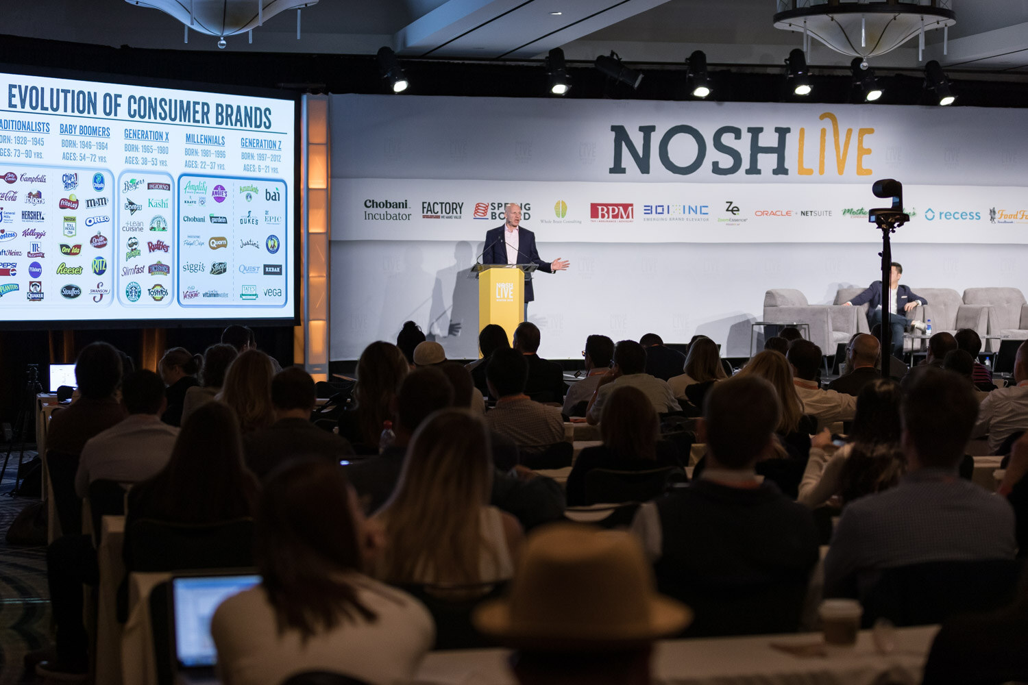 A presenter speaks from the main stage to a packed ballroom with a consumer brands slide on screen.