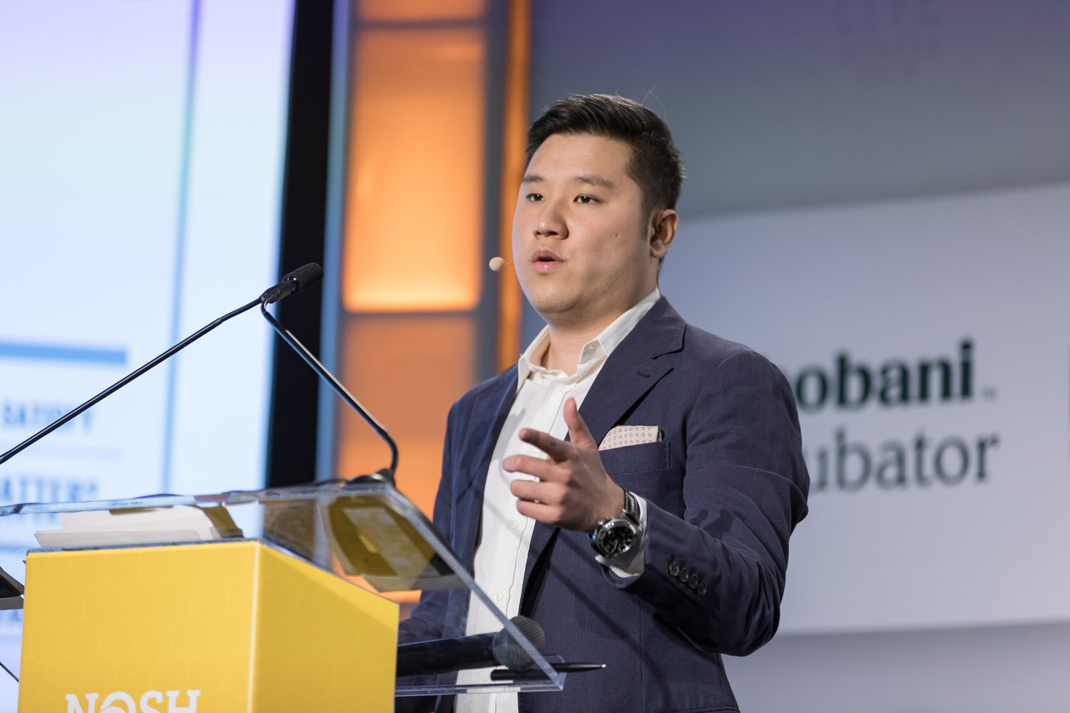 A presenter gestures with an open hand at the yellow Nosh Live lectern.