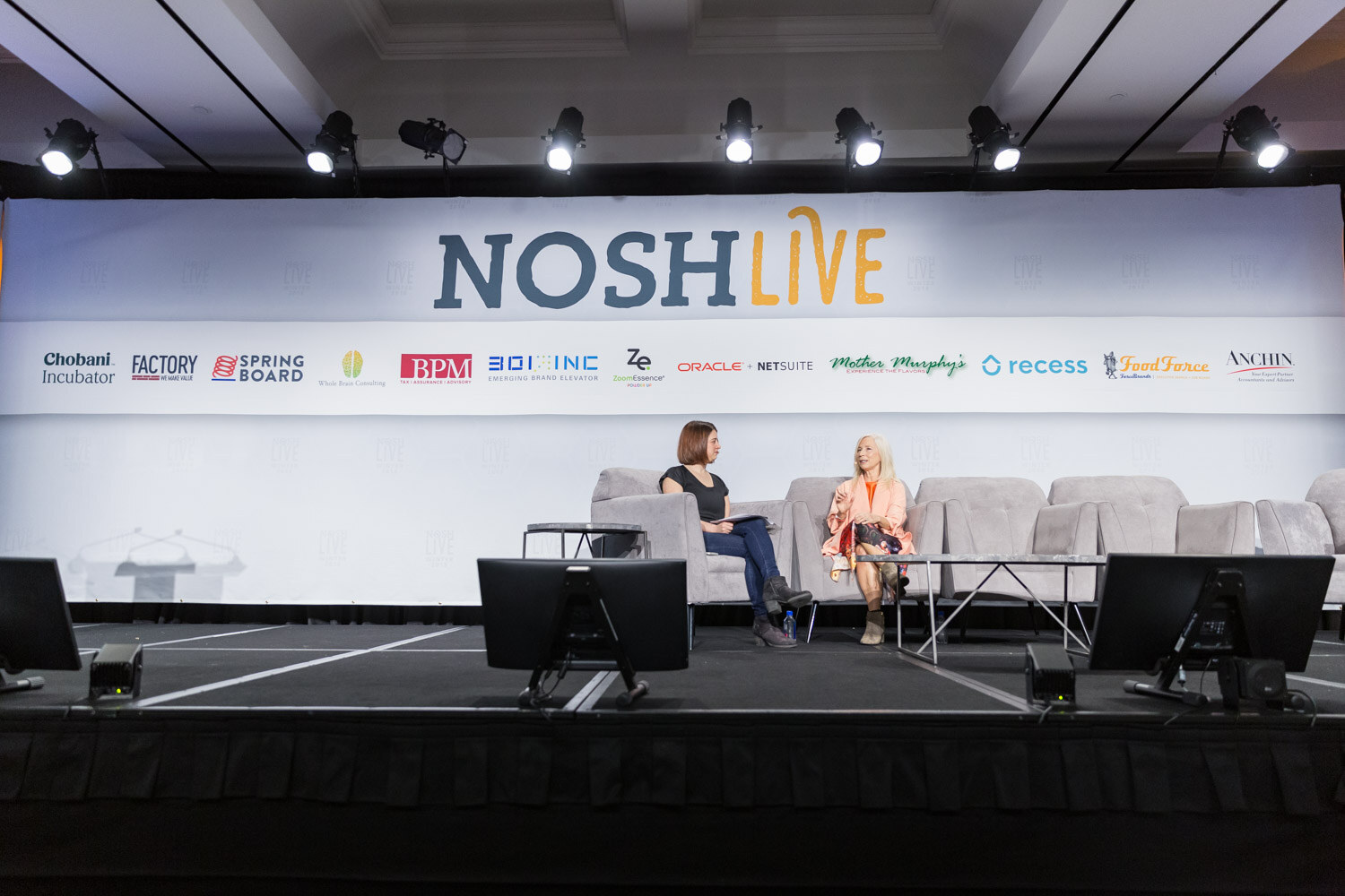 A wide view of the main stage with two women seated in gray armchairs under the Nosh Live banner.
