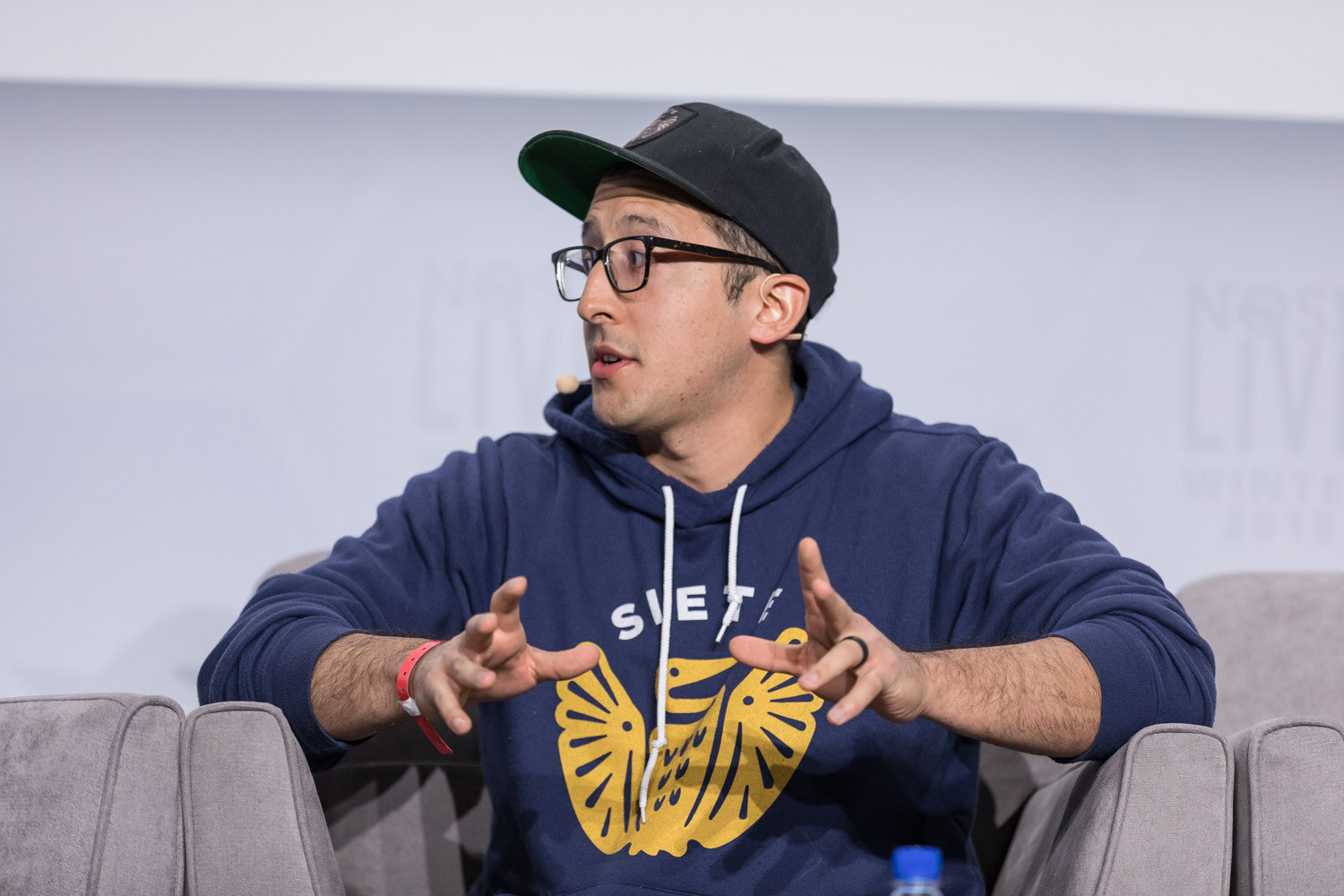 A man in a navy hoodie and cap gestures while speaking in an armchair on stage.
